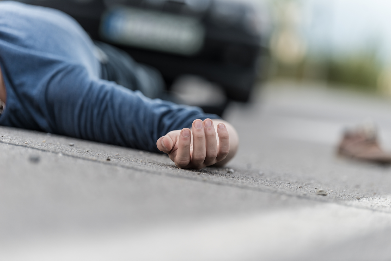 Close-up of a person's hand lying on the pavement after a pedestrian accident, with a blurred car in the background.